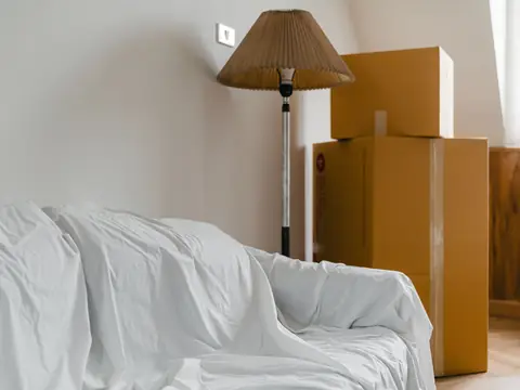 Stacked cardboard moving boxes beside a sheet-covered sofa and a lamp in a room