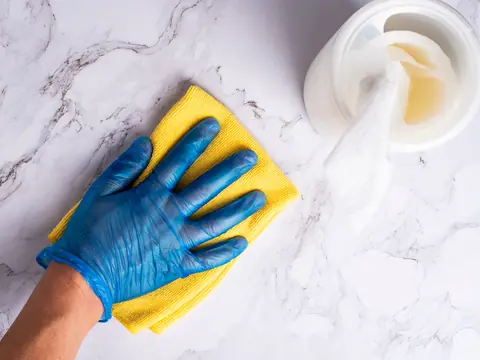 A blue-gloved hand wiping a marble countertop with a yellow cloth