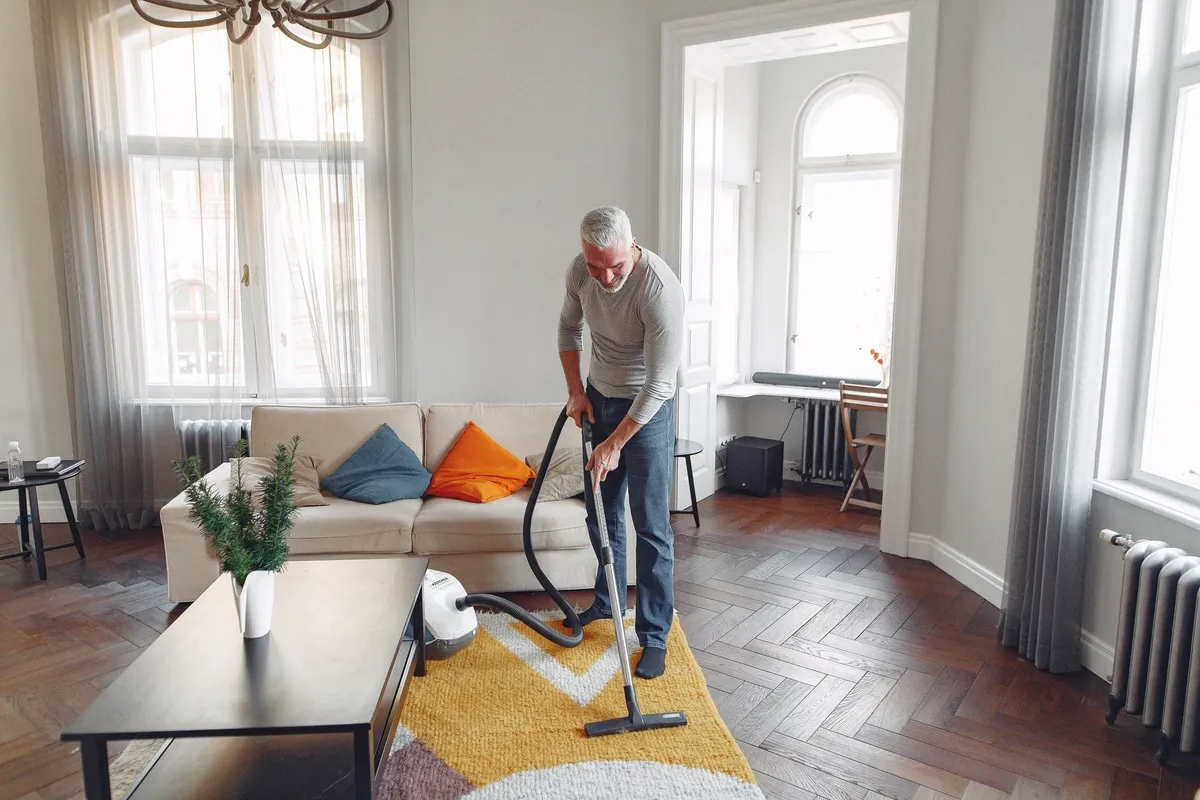 A focused grey-haired man with a beard vacuums a carpet at home, preparing for move out cleaning.
